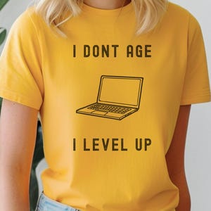 May include: A yellow t-shirt with the text "I don't age I level up" and a black and white illustration of a laptop computer.