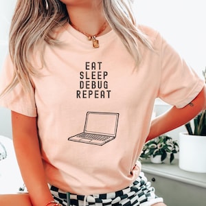 Pode incluir: Uma camiseta rosa com o texto "Eat Sleep Debug Repeat" e um gráfico de um laptop. A pessoa que está usando a camiseta está sentada em uma superfície branca e felpuda e segurando uma xícara de café.