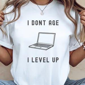 Könnte beinhalten: Weißes T-Shirt mit einer schwarzen Grafik eines Laptops und dem Text "I don't age I level up".