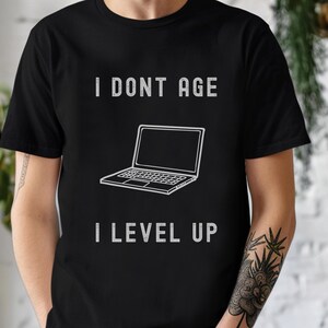 Könnte beinhalten: Schwarzes T-Shirt mit weißem Text, der "Ich werde nicht älter, ich level up" lautet, und einer Grafik eines Laptops.
