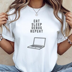 Pode incluir: Camiseta branca com um gráfico preto de um laptop e o texto "Eat Sleep Debug Repeat".