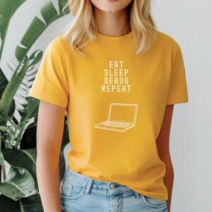 May include: Mustard yellow t-shirt with the text "EAT SLEEP DEBUG REPEAT" and a line drawing of a laptop. The shirt is paired with blue jeans. The overall theme is tech and coding.