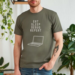 May include: Olive green t-shirt with the text "EAT SLEEP DEBUG REPEAT" and a laptop graphic in white. The shirt is a short-sleeved crew neck.