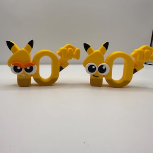 Puede incluir: Dos figuras amarillas de dibujos animados con el número "0" con rasgos de Pikachu. Cada una tiene orejas con puntas negras, una cola y una cara. Una tiene una expresión enfadada, la otra sonríe. Las figuras están sobre una superficie blanca.