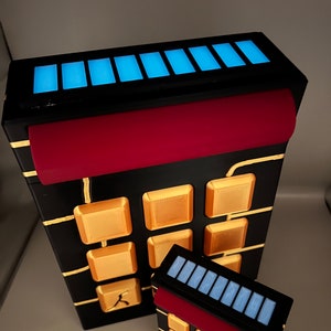 Puede incluir: Dos calculadoras negras y doradas con detalles rojos y pantallas LED azules. La calculadora más grande tiene una fila de botones dorados y la calculadora más pequeña tiene una fila de botones negros.