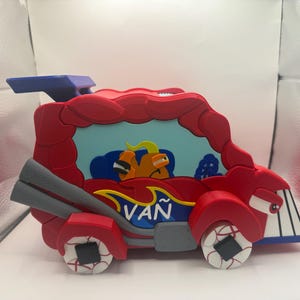 Puede incluir: Una camioneta de juguete roja con techo azul y ruedas grises. La camioneta tiene un personaje de dibujos animados en el interior y la palabra "VAN" escrita en el lateral.