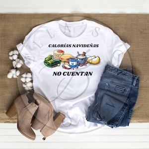 May include: White t-shirt with a graphic of Christmas food and the text "Calorías Navideñas No Cuentan". The graphic includes tamales, a cake, a cup of coffee, a bowl of hot chocolate, and a pastry. The t-shirt is paired with blue jeans and brown boots.