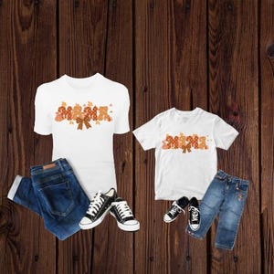 Pode incluir: Duas camisetas brancas com as palavras "Mama" e "Mema" em letras de outono laranja e marrom. As camisetas são combinadas com jeans azul e tênis.