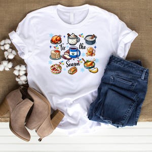 May include: White t-shirt with a festive design featuring various holiday foods and the text "Tis the Season".