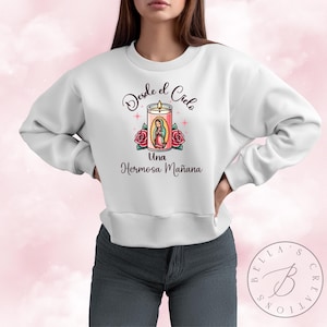 May include: White sweatshirt with a graphic of a candle with a religious figure inside. The text on the sweatshirt reads "Desde el Cielo Una Hermosa Mañana".