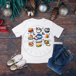 May include: White t-shirt with a festive design featuring various holiday foods and the text "Tis the Season".