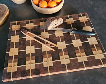 May include: A wooden cutting board with a checkered pattern in brown and light wood. A knife and tongs are on the board, along with a piece of bread.