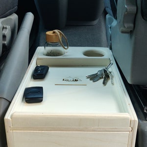 Peut inclure: Une boîte de rangement en bois clair avec un couvercle, conçue pour un van. La boîte comporte deux compartiments et un petit porte-gobelet. La boîte est gravée au laser avec un motif de montagnes et le texte "VANLIFE".
