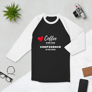 Puede incluir: Camiseta de béisbol de color blanco y negro con un corazón rojo y el texto "Coffee in one hand, Confidence in the other".