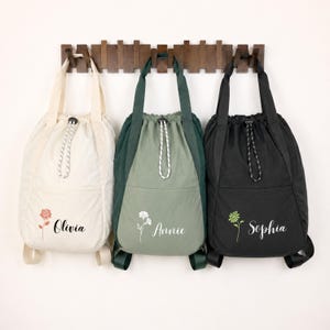 May include: Three drawstring backpacks in cream, sage green, and black. Each bag features a top handle, a drawstring closure with a cord, and a front pocket. The bags are personalized with floral illustrations and the names Olivia, Annie, and Sophia.