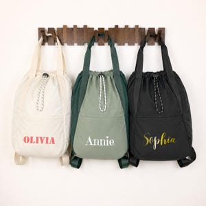Peut inclure: Trois sacs à dos à cordon suspendus sur un support en bois. Le sac de gauche est crème avec le nom OLIVIA en rouge. Le sac central est vert sauge avec le nom Annie en blanc. Le sac de droite est noir avec le nom Sophia en écriture dorée.