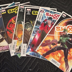 Puede incluir: Una pila de cómics con el título "S.W.O.R.D." y varios personajes de Marvel. Las portadas muestran ilustraciones vibrantes con escenas de acción y el logotipo de Marvel. Los cómics están apilados sobre una superficie oscura.