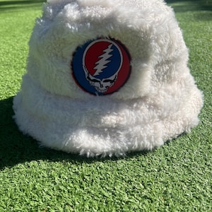 May include: A white, fluffy bucket hat with a round patch featuring a red, blue, and white Steal Your Face logo. The hat is made of a soft, textured material and sits on a green surface.