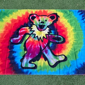 May include: A tie-dye flag featuring a black and white outline of a dancing bear. The bear is wearing a yellow starburst collar. The flag is a vibrant mix of red, orange, yellow, green, blue, and purple.