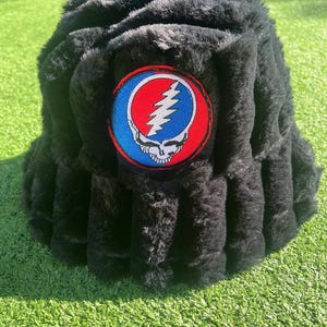 May include: Black faux fur bucket hat with a colorful patch featuring a skull, lightning bolt, and a red, white, and blue color scheme. The hat is sitting on green grass.