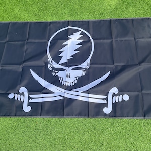 Puede incluir: Bandera negra con una calavera blanca y un rayo en la frente, cruzada por dos espadas blancas. La bandera tiene un borde blanco y ojales para colgarla. Un diseño único para los fanáticos de la música y los piratas.