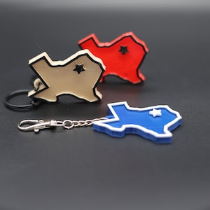 May include: Three Texas-shaped keychains, one red, one beige, and one blue. Each keychain has a silver chain and a silver clasp. The red and beige keychains have a black star in the center of the state shape. The blue keychain has a white star in the center of the state shape.