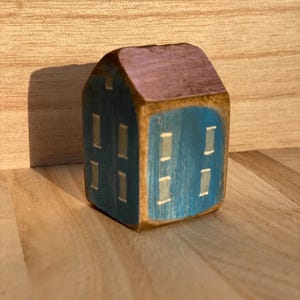 Puede incluir: Una peque&ntilde;a figura de casa de madera pintada a mano con una fachada azul oscuro, ventanas rectangulares claras y un techo de tonos morados. La veta natural de la madera es visible en los bordes del bloque, creando una estructura s&oacute;lida.