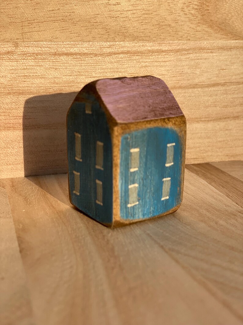 Puede incluir: Una peque&ntilde;a figura de casa de madera pintada a mano con una fachada azul oscuro, ventanas rectangulares claras y un techo de tonos morados. La veta natural de la madera es visible en los bordes del bloque, creando una estructura s&oacute;lida.