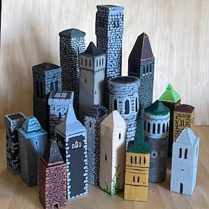 Puede incluir: Una colección de torres y edificios de madera en miniatura pintados en varios colores, incluyendo gris, azul, amarillo y marrón. Las estructuras presentan patrones detallados como ladrillos, ventanas y techos puntiagudos, dispuestos como una pequeña ciudad.