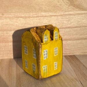 Puede incluir: Una peque&ntilde;a figura de casa de madera pintada a mano en un color amarillo vibrante. La estructura cuenta con dos ventanas abuhardilladas en el techo y varias ventanas rectangulares pintadas en blanco en las fachadas frontal y lateral.
