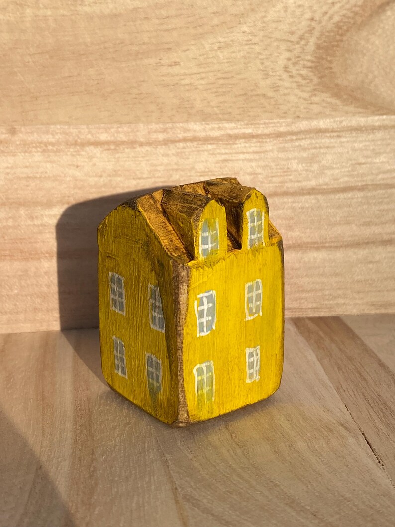 Puede incluir: Una peque&ntilde;a figura de casa de madera pintada a mano en un color amarillo vibrante. La estructura cuenta con dos ventanas abuhardilladas en el techo y varias ventanas rectangulares pintadas en blanco en las fachadas frontal y lateral.
