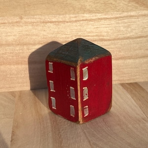 Puede incluir: Un peque&ntilde;o bloque de madera pintado a mano con forma de casa, con exterior rojo y techo azul oscuro. La estructura presenta ventanas rectangulares blancas pintadas en dos lados visibles, con un acabado desgastado que muestra la veta de la madera.