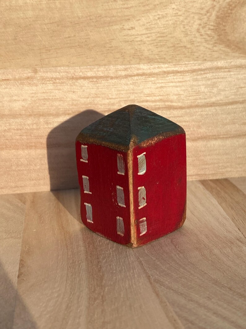 Puede incluir: Un peque&ntilde;o bloque de madera pintado a mano con forma de casa, con exterior rojo y techo azul oscuro. La estructura presenta ventanas rectangulares blancas pintadas en dos lados visibles, con un acabado desgastado que muestra la veta de la madera.