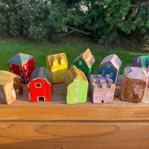 Puede incluir: Una colecci&oacute;n de once peque&ntilde;as figuras de casas de madera pintadas a mano en varios colores, incluyendo rojo, amarillo, verde, azul, morado y marr&oacute;n. Cada edificio en miniatura presenta ventanas y puertas pintadas con un acabado r&uacute;stico y desgastado.
