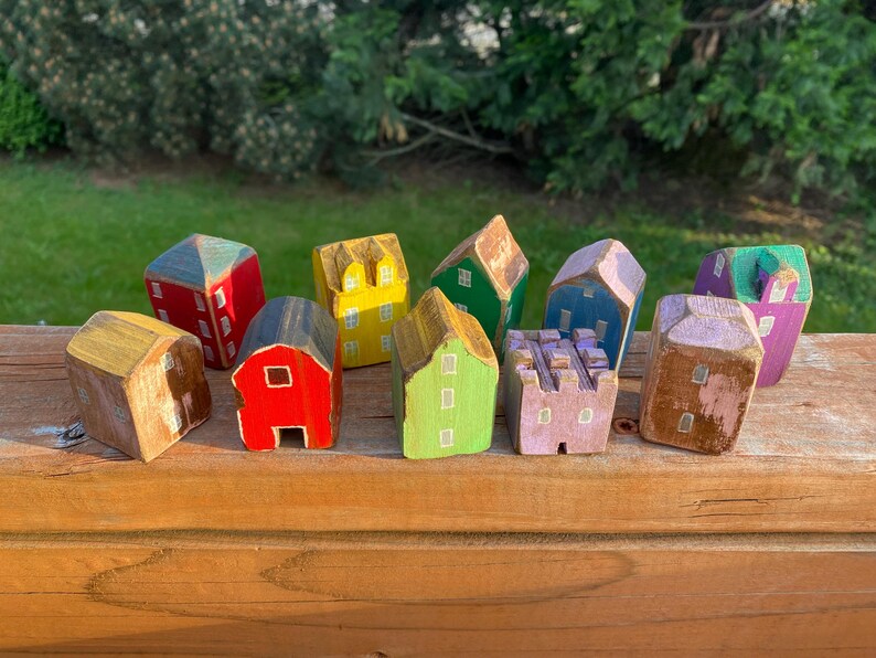 Puede incluir: Una colecci&oacute;n de once peque&ntilde;as figuras de casas de madera pintadas a mano en varios colores, incluyendo rojo, amarillo, verde, azul, morado y marr&oacute;n. Cada edificio en miniatura presenta ventanas y puertas pintadas con un acabado r&uacute;stico y desgastado.