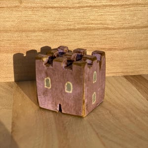 Puede incluir: Una peque&ntilde;a figura de castillo de madera pintada a mano en un color malva apagado. La estructura presenta una parte superior almenada, una base cuadrada y varias ventanas peque&ntilde;as y rectangulares pintadas en verde claro p&aacute;lido.