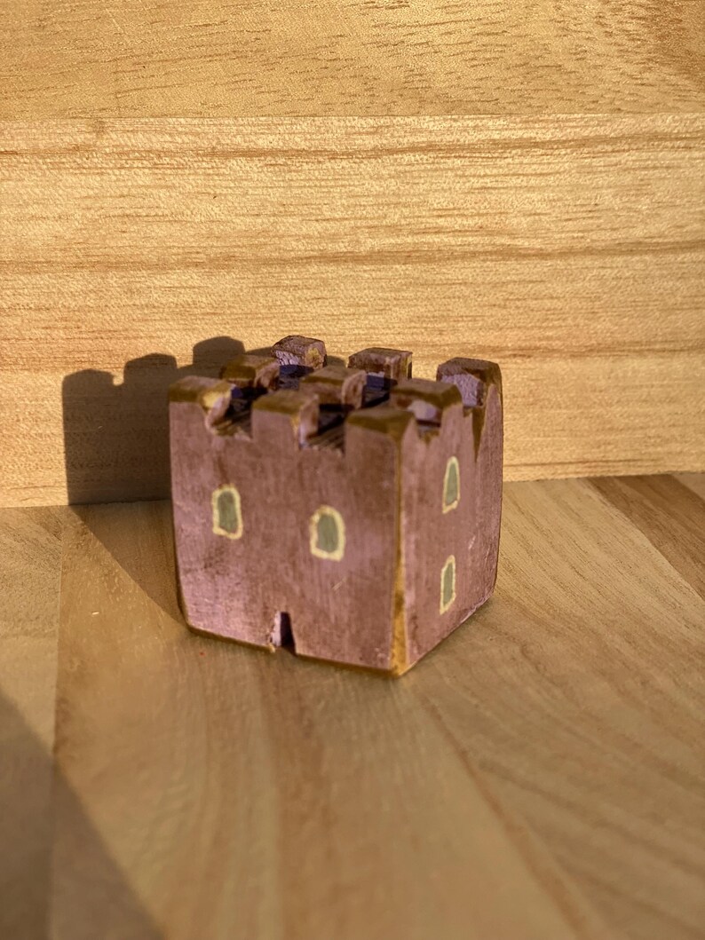 Puede incluir: Una peque&ntilde;a figura de castillo de madera pintada a mano en un color malva apagado. La estructura presenta una parte superior almenada, una base cuadrada y varias ventanas peque&ntilde;as y rectangulares pintadas en verde claro p&aacute;lido.