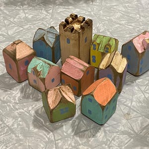 Puede incluir: Una colección de diez pequeñas figuras de casas de madera pintadas a mano y una torre de castillo central. Cada pieza presenta un acabado desgastado con colores pastel como rosa, azul, verde, amarillo y naranja, con ventanas azules pintadas.