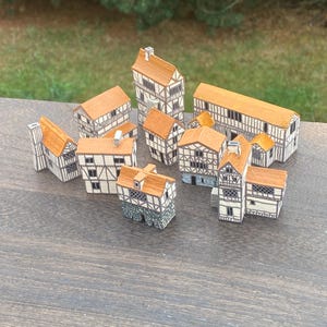 Op de afbeelding: Een verzameling miniatuurmodelhuizen met bruine daken en wit en bruin vakwerk. De huizen variëren in grootte en vorm, sommige met schoorstenen en meerdere verdiepingen. De modellen zijn gerangschikt op een houten oppervlak.