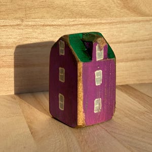 Puede incluir: Una peque&ntilde;a figura de casa de madera pintada a mano con una fachada morada y un techo verde. La estructura tiene un remate en forma de hastial y est&aacute; decorada con varias ventanas blancas rectangulares pintadas en las superficies.