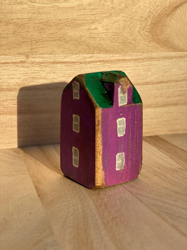 Puede incluir: Una peque&ntilde;a figura de casa de madera pintada a mano con una fachada morada y un techo verde. La estructura tiene un remate en forma de hastial y est&aacute; decorada con varias ventanas blancas rectangulares pintadas en las superficies.