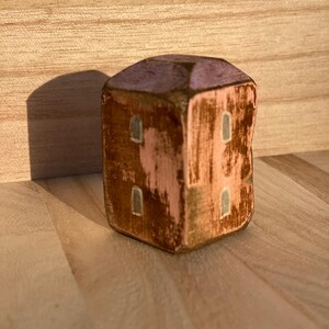 Puede incluir: Una peque&ntilde;a figura de casa de madera en forma de bloque con un acabado desgastado. La estructura presenta un techo de color violeta claro, paredes rosas con parches marrones desgastados y cuatro ventanas rectangulares pintadas en gris claro.