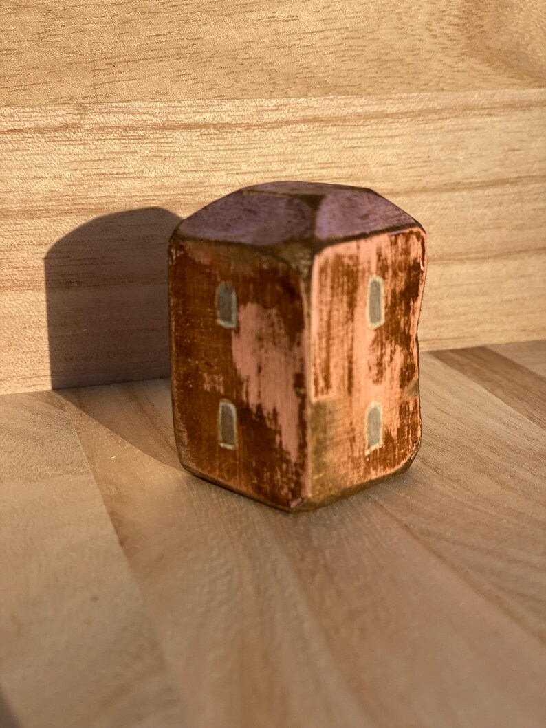 Puede incluir: Una peque&ntilde;a figura de casa de madera en forma de bloque con un acabado desgastado. La estructura presenta un techo de color violeta claro, paredes rosas con parches marrones desgastados y cuatro ventanas rectangulares pintadas en gris claro.