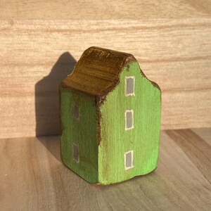 Puede incluir: Una peque&ntilde;a figura de casa de madera pintada de color verde brillante con un techo marr&oacute;n oscuro. La fachada frontal presenta tres ventanas rectangulares pintadas en gris claro con bordes blancos, y el lateral muestra dos ventanas adicionales.