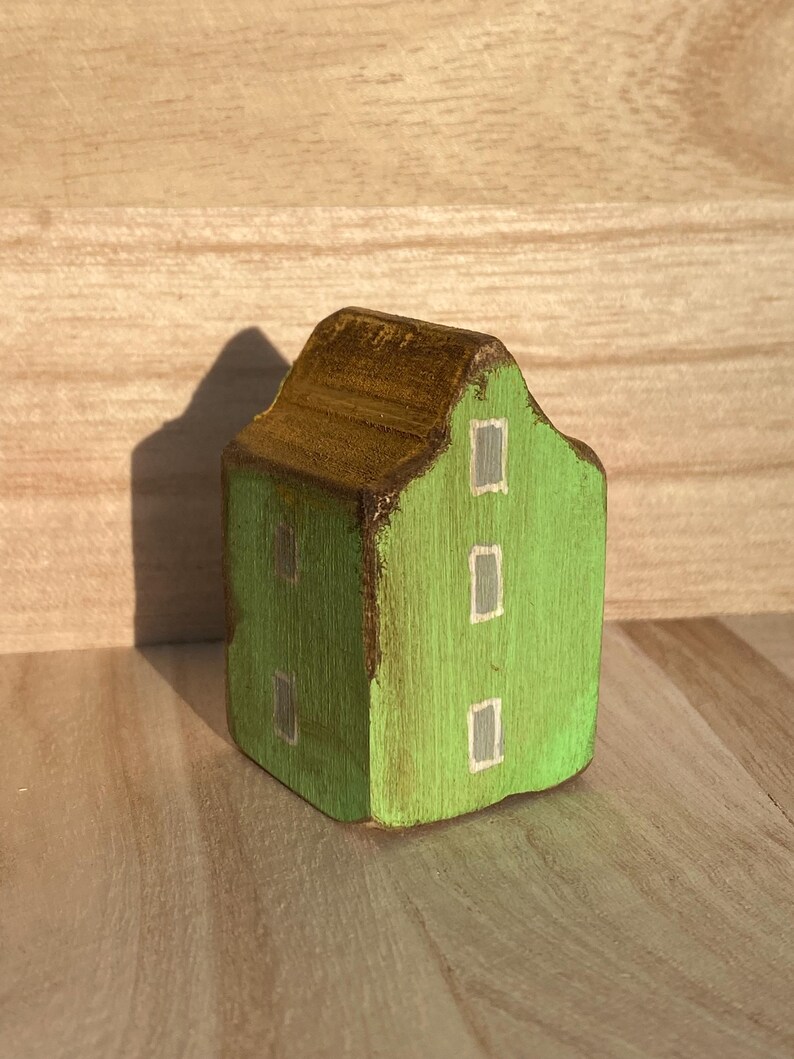 Puede incluir: Una peque&ntilde;a figura de casa de madera pintada de color verde brillante con un techo marr&oacute;n oscuro. La fachada frontal presenta tres ventanas rectangulares pintadas en gris claro con bordes blancos, y el lateral muestra dos ventanas adicionales.