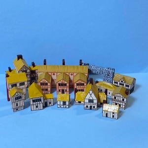 Puede incluir: Una colección de edificios de madera en miniatura con diseño estilo Tudor, paredes blancas, entramado de madera marrón oscuro y techos amarillos. El conjunto incluye varias estructuras de casas y un puente con patrón de piedra sobre fondo azul.