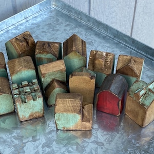Puede incluir: Una colección de pequeñas figuras de casas de madera talladas a mano con un acabado desgastado en tonos de madera natural y parches de pintura verde azulada. Una casa tiene el frente rojo y otra presenta un borde superior almenado.