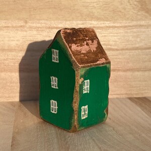 Puede incluir: Una peque&ntilde;a figura de casa de madera pintada en un color verde vibrante con un techo marr&oacute;n. La estructura presenta ventanas blancas pintadas a mano en las fachadas frontal y lateral, con textura de veta de madera visible y bordes desgastados.