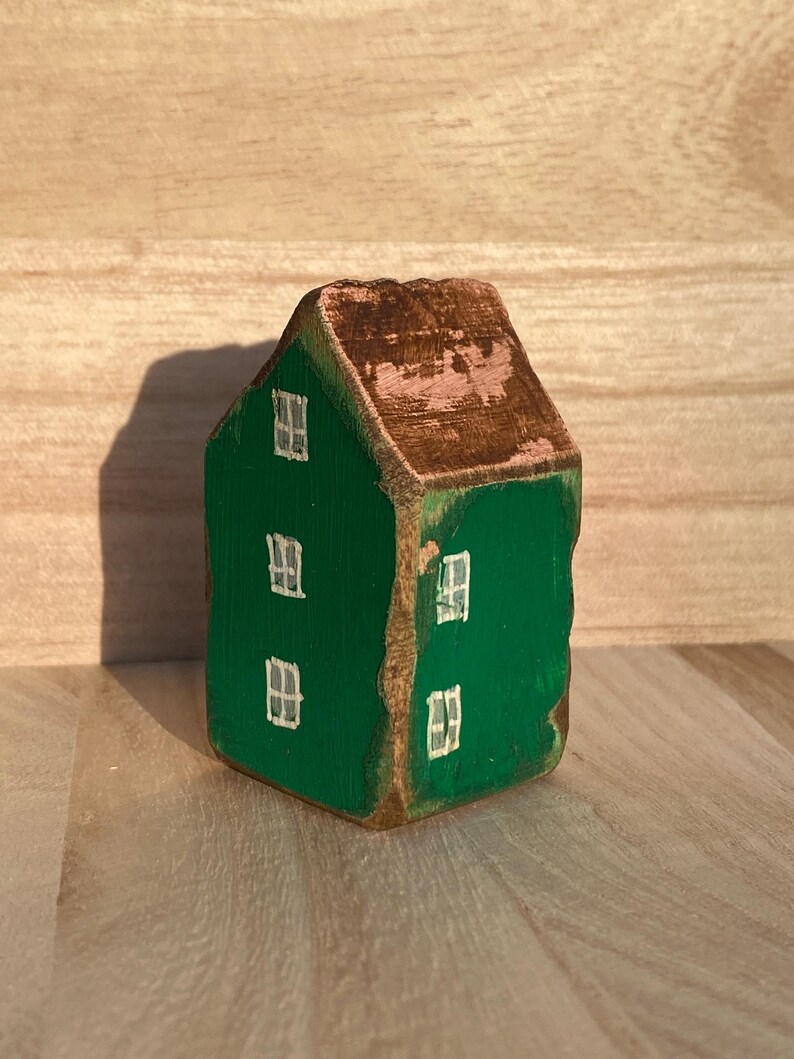Puede incluir: Una peque&ntilde;a figura de casa de madera pintada en un color verde vibrante con un techo marr&oacute;n. La estructura presenta ventanas blancas pintadas a mano en las fachadas frontal y lateral, con textura de veta de madera visible y bordes desgastados.