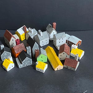 May include: A collection of small, hand-painted wooden house figurines in various architectural styles. The miniature buildings feature diverse roof colors including red, brown, yellow, and gray, with painted windows and chimneys on many structures.
